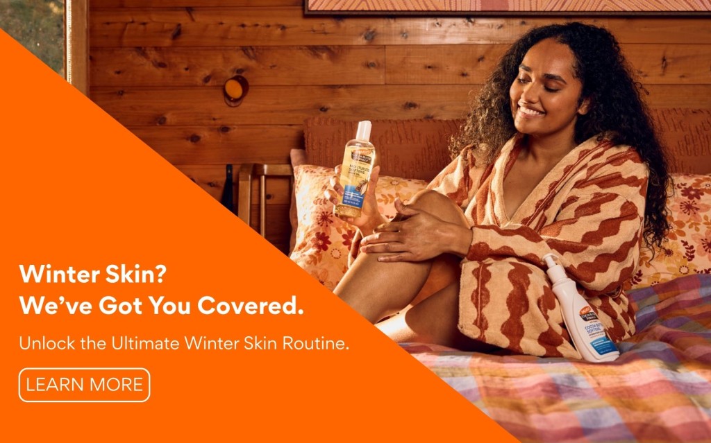 Unlock the Ultimate Winter Skin Routine with Palmer's Cocoa Butter - A smiling women shown applying Palmer's Cocoa Butter Body Oil to legs whilst on a bed in a fluffy robe. 