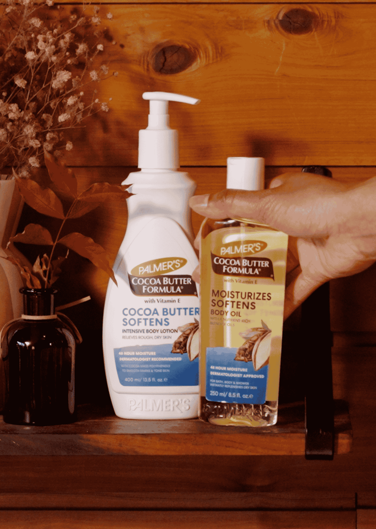 Palmer's Cocoa Butter Body Lotion and Body Oil shown on a wooden shelf and a hand is reaching to pick up the oil