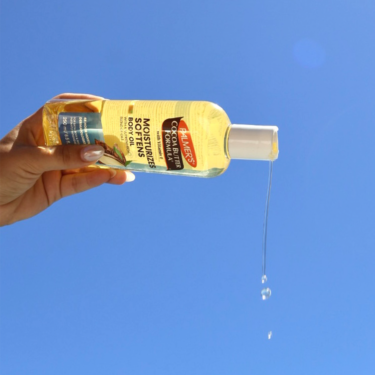 Hand holding a bottle of Palmer's Cocoa Butter Formula body oil against a blue sky.
