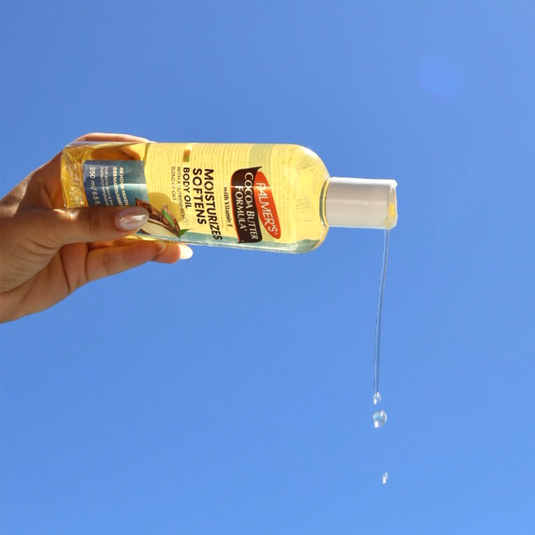 Hand holding a bottle of Palmer's Cocoa Butter Formula body oil against a blue sky.