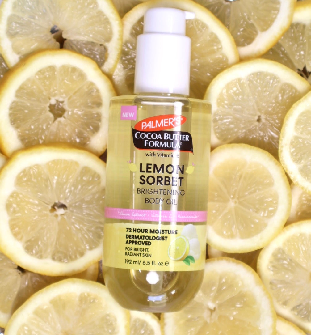 A bottle of Palmer Lemon Sorbet Brightening Body Oil centered against a background of sliced lemon wedges. The bottle has a white pump and a yellow-tinted liquid inside, with a label featuring pink and green accents.