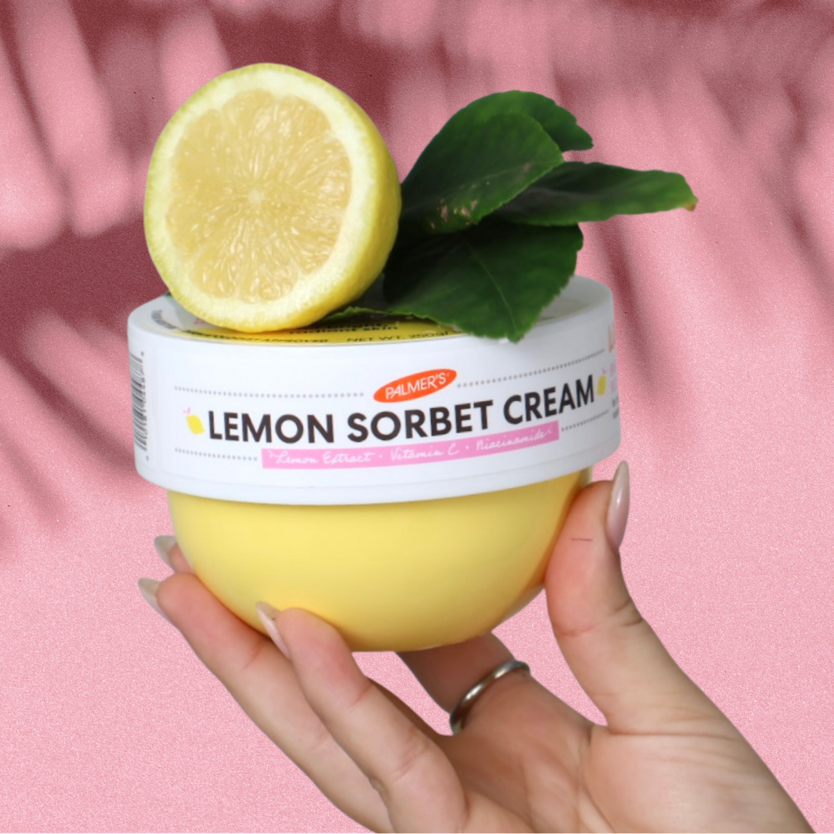 A hand holding a jar of Pampas Lemon Sorbet Cream, topped with a fresh lemon half and green leaves, set against a pink background with leaf shadows.