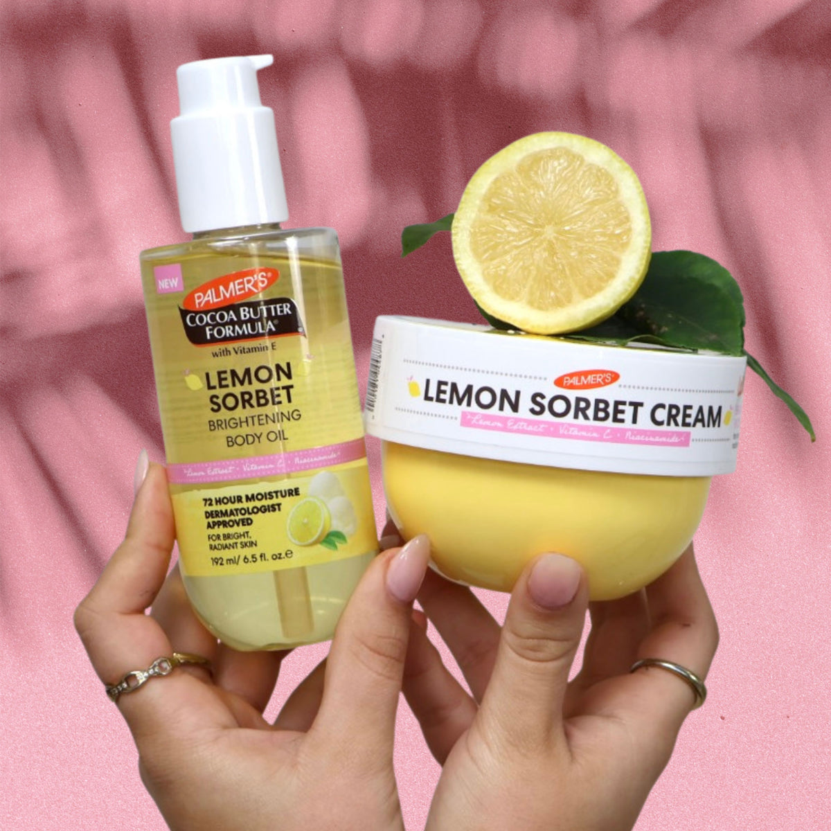 Two hands holding Palmer's Lemon Sorbet body care products against a pink fabric background: a bottle of Lemon Sorbet Brightening Body Oil on the left and a jar of Lemon Sorbet Cream on the right with a lemon slice on top.