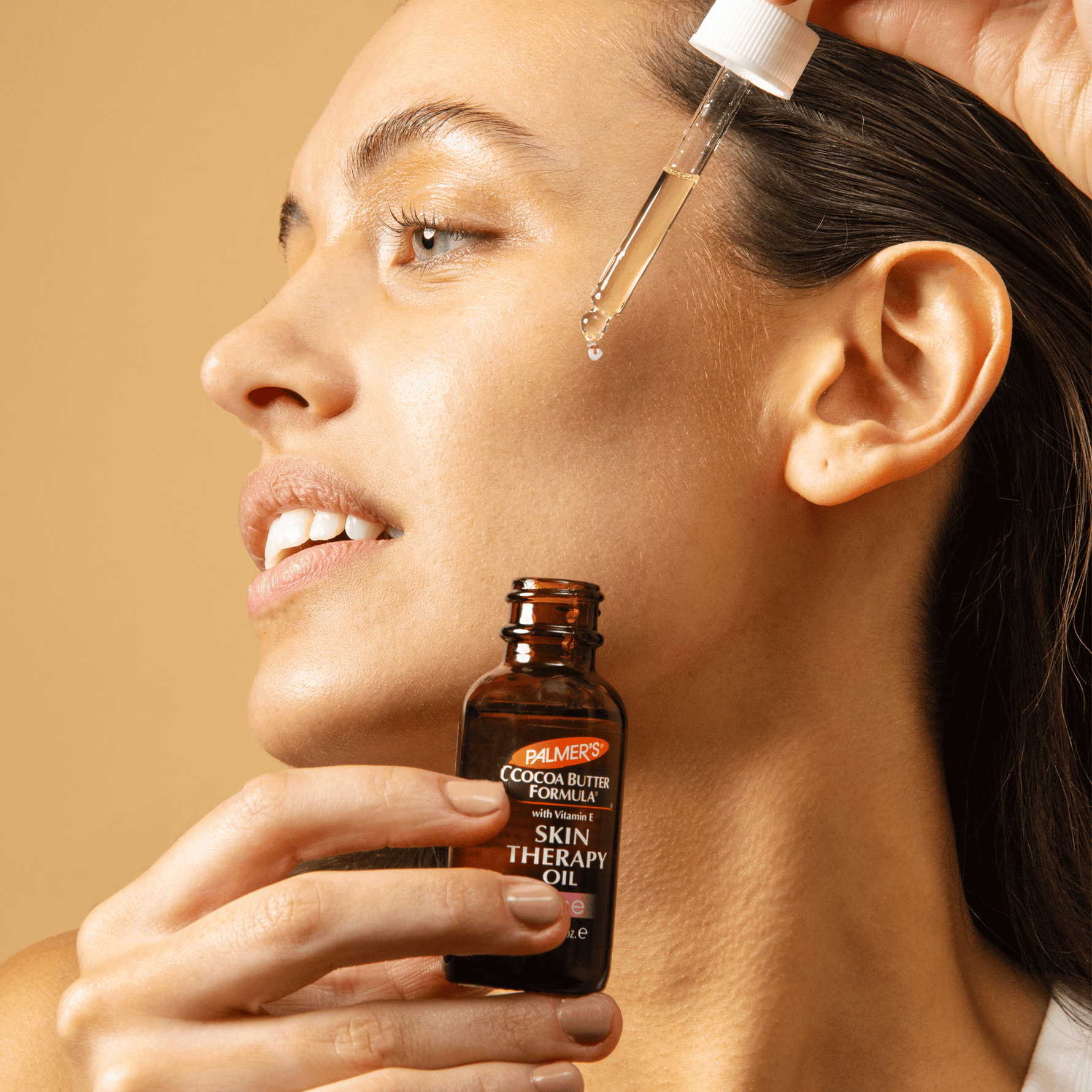 Woman applying a dropper of oil to her face with a bottle of Cocoa Butter Formula Skin Therapy Oil in front.